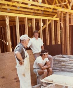 Photograph of two men standing and another man sitting in a partially-framed house.